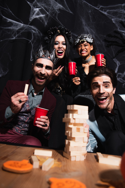 opgewonden multi-etnische vrienden in halloween kostuums op zoek naar camera in de buurt van houten blokken spel op zwart  - Foto, afbeelding