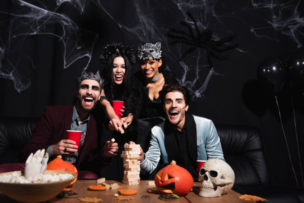 возбужденные мультикультурные женщины в костюмах Хэллоуина играть деревянные блоки игра с мужчинами на черном  - Фото, изображение