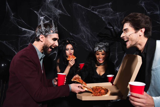 happy man in vampire king crown taking pizza near multiethnic friends on black - Photo, Image