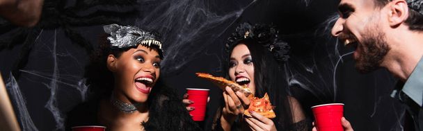 emocionado Africano ameican mulher segurando pizza durante halloween festa com amigos multiétnicos em preto, banner - Foto, Imagem