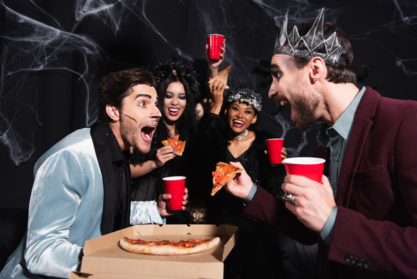 Erstaunte Männer, die ihren Freund in der Nähe einer Pizza anschauen und aufgeregte interrassische Frauen in Halloween-Kostümen auf schwarz - Foto, Bild