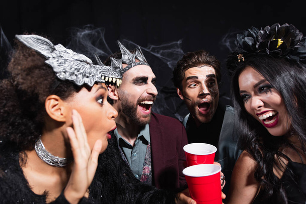 excited african american woman in wolf mask talking to amazed multiethnic friends during party on black  - Foto, Bild