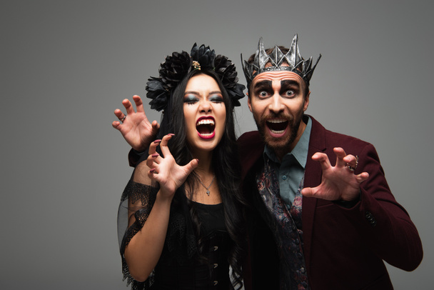 interracial pár v upíři halloween kostýmy vrčení a ukazující děsivé gesto izolované na šedé - Fotografie, Obrázek