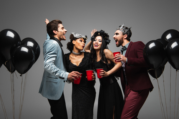 multiethnic friends with plastic cups posing near black balloons on halloween party isolated on grey - Foto, Imagen