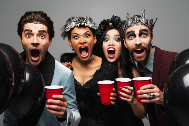 жуткие многоэтнические друзья с пластиковыми чашками, кричащие и рычащие на камеру на вечеринке Хэллоуина изолированные на сером - Фото, изображение
