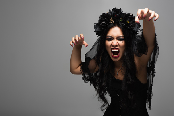 spooky asian woman in vampire halloween costume growling with closed eyes and showing scary gesture isolated on grey - Foto, Imagem