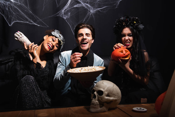 amigos multiétnicos felizes com pipoca, mão de brinquedo e abóbora halloween olhando para a câmera perto do crânio assustador no preto - Foto, Imagem