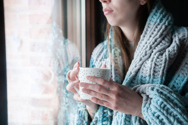 Νεαρή γυναίκα με μάλλινη κουβέρτα το χειμώνα κοιτάζοντας έξω από το παράθυρο κρατώντας ένα ζεστό ποτό - Φωτογραφία, εικόνα