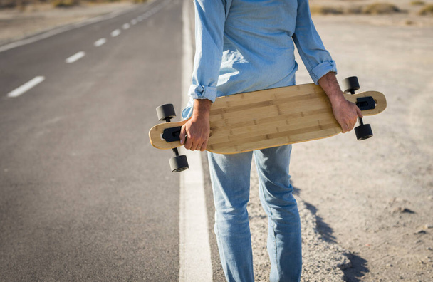 Руки людини тримають скейтборд на дорозі. Вид ззаду чоловіка в джинсовому одязі, що тримає скейтборд на сільській дорозі
. - Фото, зображення