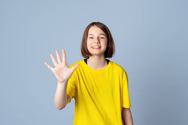 Freundliches, fröhliches Teenie-Mädchen in gelbem, lässigem T-Shirt, das mit der Handfläche wedelt, freundlich grüßt oder Hallo sagt, vor hellgrauem Hintergrund stehend. Konzept positiver Emotionen - Foto, Bild