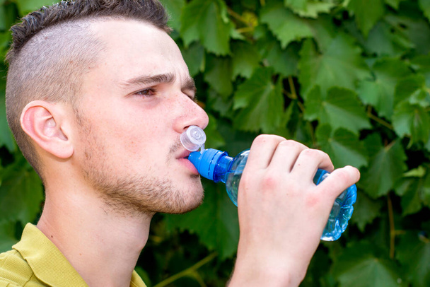 Ein junger Mann mit stylischer Frisur trinkt vor dem Hintergrund grünen Laubs Wasser aus einer Flasche. Hochwertiges Foto - Foto, Bild