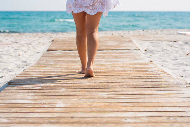 Низкая часть женщины идет по деревянной доске среди песка, ведущего к морской воде.  - Фото, изображение