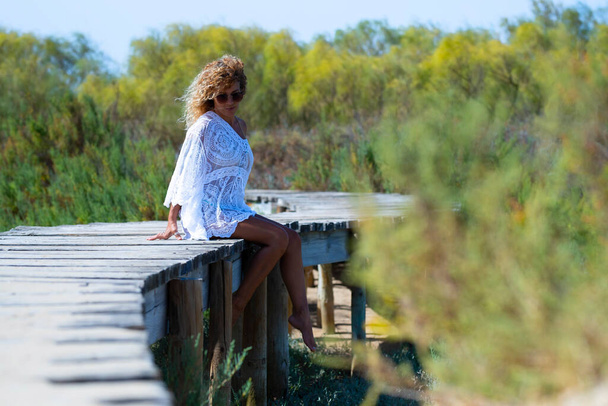 Attraktive Frau im weißen Kleid sitzt barfuß auf einer erhöhten Fußgängerbrücke, die in Richtung natürlicher Wald führt. - Foto, Bild