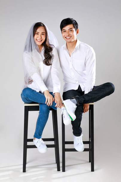 白いシャツを着た若い魅力的なアジアのカップルと、ウェディングベールを着たジーンズの女性が笑顔で座っています。結婚式前の写真のコンセプト. - 写真・画像