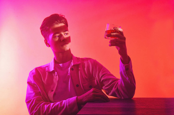 Un hombre sobre un fondo naranja-púrpura con un vaso de whisky - Foto, Imagen