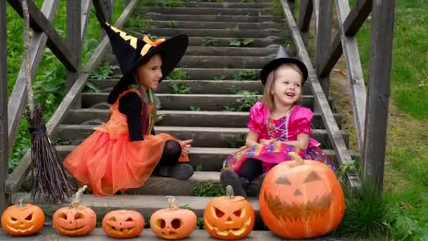 魔女の格好をした2人の小さな女の子が家の前の古いはしごの階段に座っていて、甘く笑っています。ハロウィーンの面白い笑いの女の子は階段で御馳走を待っています. - 映像、動画