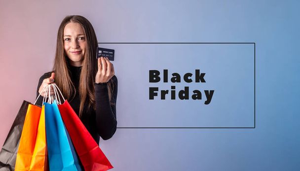 Woman in a black dress is holding a credit card and colorful paper bags with purchases from a shop in her hands on a blue pink background. Inscription Black Friday. Concept of shopping and sale. - Foto, Imagem