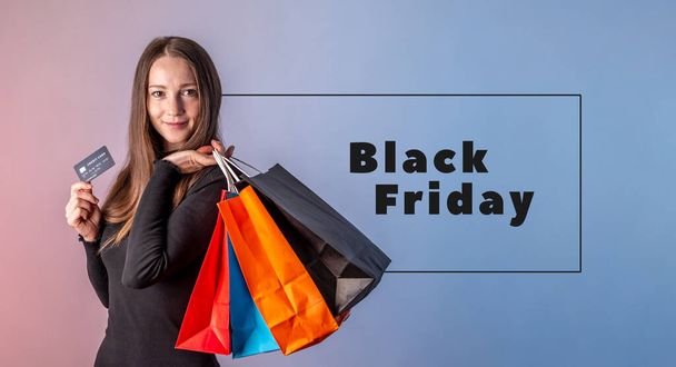 Woman in a black dress is holding a credit card and colorful paper bags with purchases from a shop in her hands on a blue pink background. Inscription Black Friday. Concept of shopping and sale. - Foto, afbeelding