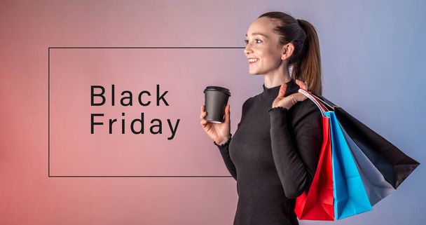 Woman in a black dress is holding a cup of coffee and colorful paper bags with purchases from a shop in her hands and smiling. Inscription Black Friday. Concept of shopping, sale and special offers. - Zdjęcie, obraz