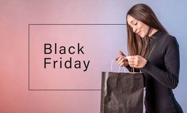 Young woman in a black dress is holding a paper bag from the shop in her hands and looking into it with a smile. Inscription is black Friday. Concept of shopping, sale and profitable purchases. - Fotoğraf, Görsel