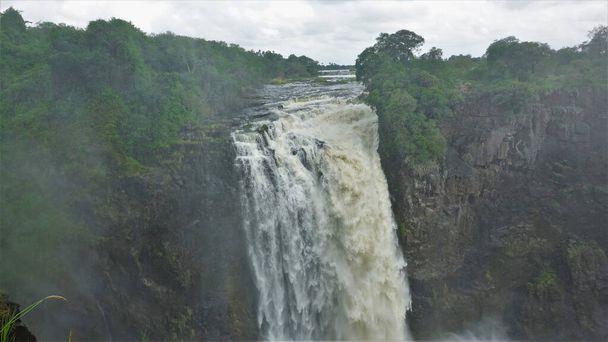 Река Замбези течёт от края плато в ущелье, образуя водопад Виктория. Вокруг пышная зеленая растительность. Туман висит над пропастью. Зимбабве - Фото, изображение