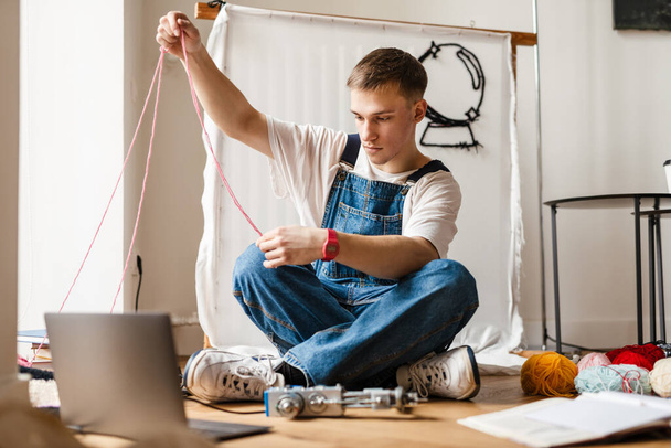 Mladý bílý muž pomocí notebooku a šicí stroj při práci na řemeslné koberec doma - Fotografie, Obrázek