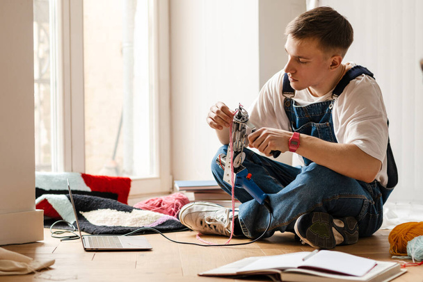 Young white man using laptop and sewing machine while working on craft rug at home - Fotografie, Obrázek