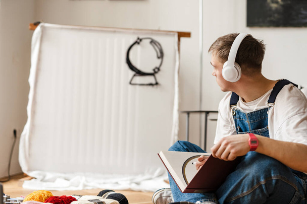 Fiatal férfi fülhallgatóban jegyzetel, miközben otthon kézműves szőnyegen dolgozik. - Fotó, kép