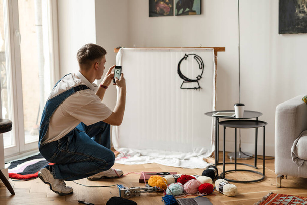 Young white man taking photo on cellphone while working on craft rug at home - Fotoğraf, Görsel