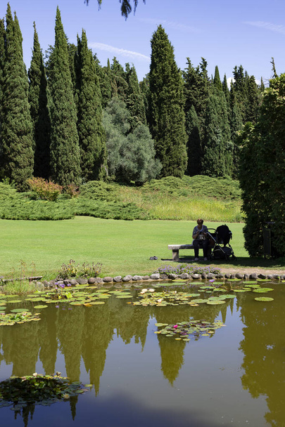 Grootmoeder met kinderwagen zittend bij een kleine vijver op een zonnige dag. Sigurt Garden Park, Valeggio sul Mincio, Veneto, Italië. Verticaal schot. - Foto, afbeelding