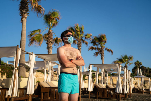 Muž v lékařské masce stojí sám na prázdné pláži v blízkosti lehátka a palmy na Kypru, Paphos město během karantény a uzamčení koronaviru. Nový normální čas dovolené po pandemii COVID-19. - Fotografie, Obrázek
