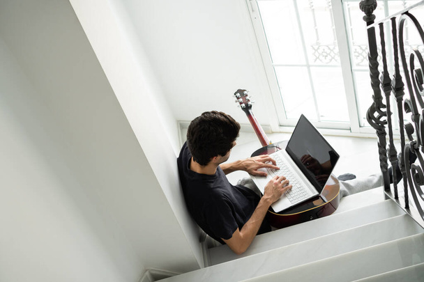 Casual άνθρωπος κάνει ένα διάλειμμα από το να παίζει κιθάρα για να χρησιμοποιήσετε ένα φορητό υπολογιστή στο σπίτι. - Φωτογραφία, εικόνα