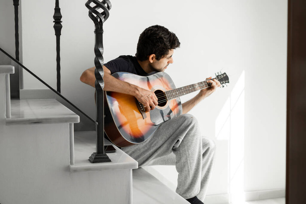 Зверху молодий чоловік грає на акустичній гітарі, сидячи на самоті на сходах біля вікна в будинку
. - Фото, зображення