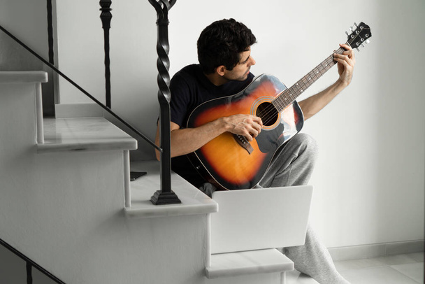 Man playing guitar using laptop for learning on staircase at home. - 写真・画像
