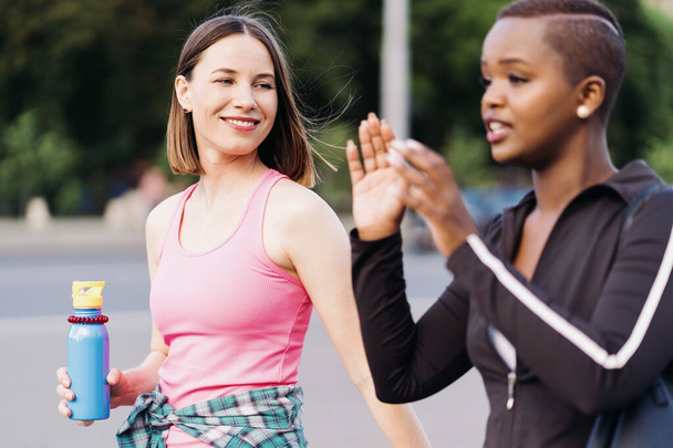 Dos chicas multiétnicas caminando para una sesión de ejercicio, comunicándose al aire libre. - Foto, imagen
