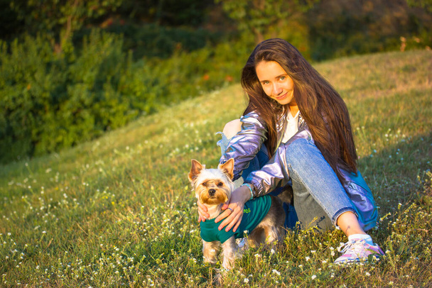 A young beautiful long-haired stylish girl in a silver raincoat, blue jeans is sitting on a park lawn, blossoming meadow hugging a Yorkshire terrier small dog, puppy. People and pets, animals lover.  - Foto, immagini