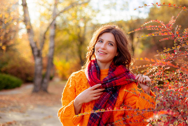 een jonge vrouw in een oranje trui wikkelt zich in een sjaal in een herfstpark - Foto, afbeelding