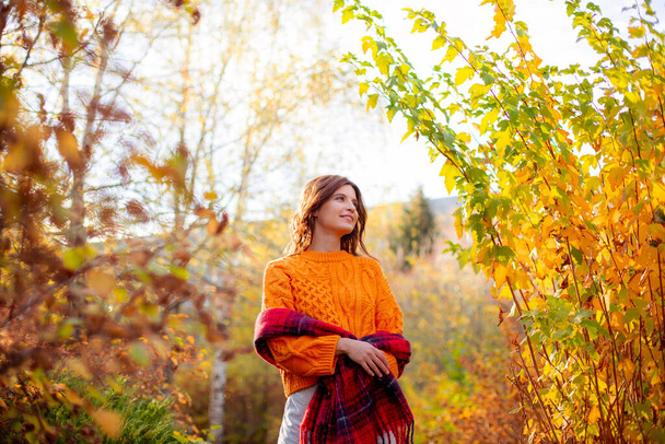 una giovane donna in un maglione arancione si avvolge in una sciarpa in un parco autunnale - Foto, immagini