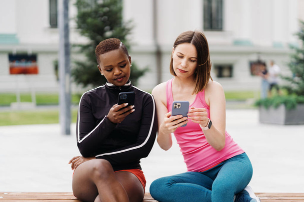 Twee multi-etnische meisjes ontspannen na de training sessie, buiten communiceren en het gebruik van de smartphone - Foto, afbeelding