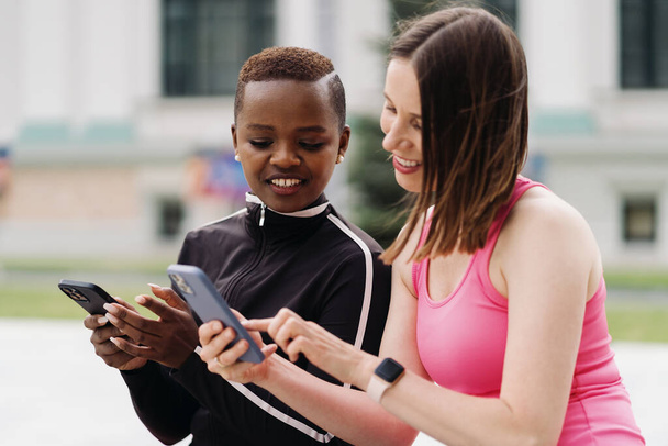 Dos chicas multiétnicas relajándose después de una sesión de ejercicio, comunicándose al aire libre y utilizando un teléfono inteligente - Foto, Imagen