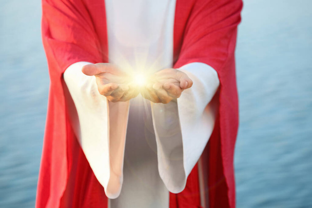 Jesus Christ near water outdoors, closeup. Miraculous light in hands - Photo, Image
