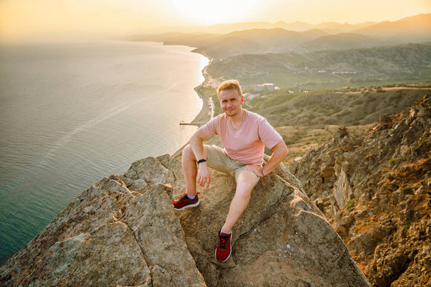 Счастливый человек сидит с распростертыми руками на краю скалы над морем с прекрасным видом на горы и село в Крыму. Завоевание высот и понятие свободы - Фото, изображение