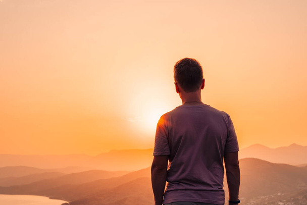 Egy fiatalember áll egy szikla szélén naplementekor, hihetetlen kilátással a hegyekre. - Fotó, kép