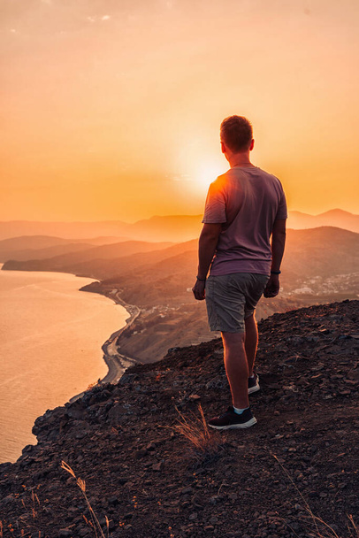 Ένας νεαρός άνδρας στέκεται στην άκρη ενός γκρεμού το ηλιοβασίλεμα με απίστευτη θέα στα βουνά. - Φωτογραφία, εικόνα