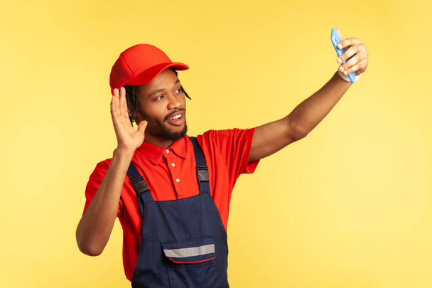 Smiling friendly workman wearing blue overalls taking selfie or having video call with client, waving hand, saying hello, being ready to execute order. Indoor studio shot isolated on yellow background - Foto, Bild