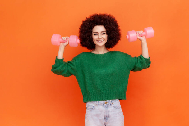 Positive woman with Afro hairstyle wearing green casual style sweater raising arms with pink dumbbells, training biceps and triceps, healthy lifestyle. Indoor studio shot isolated on orange background - Фото, изображение