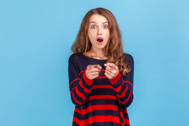 Astonished woman with wavy hair wearing striped casual style sweater pointing to camera with surprised shocked expression, making choice. Indoor studio shot isolated on blue background. - Foto, Bild