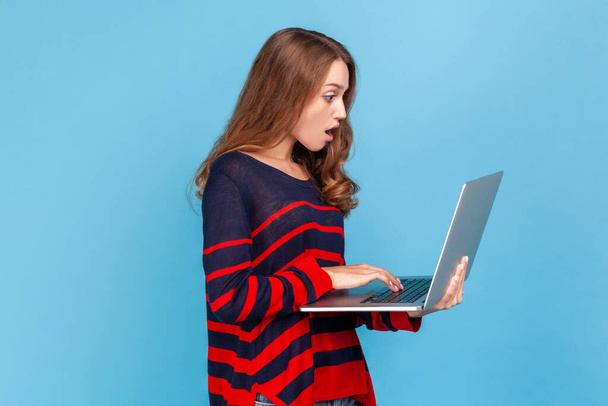 Side view portrait of astonished woman with wavy hair in striped casual style sweater working online, looking at laptop display with shocked expression. Indoor studio shot isolated on blue background. - Фото, изображение