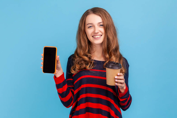 Good looking woman with wavy hair wearing striped casual style sweater standing holding coffee to go and smart phone with empty black screen. Indoor studio shot isolated on blue background. - Fotografie, Obrázek