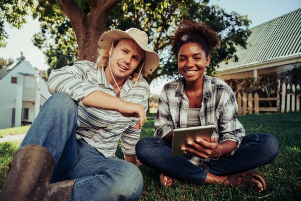 Mixed race couple smiling while enjoying time together sitting on grass scrolling through images on digital device  - Photo, image